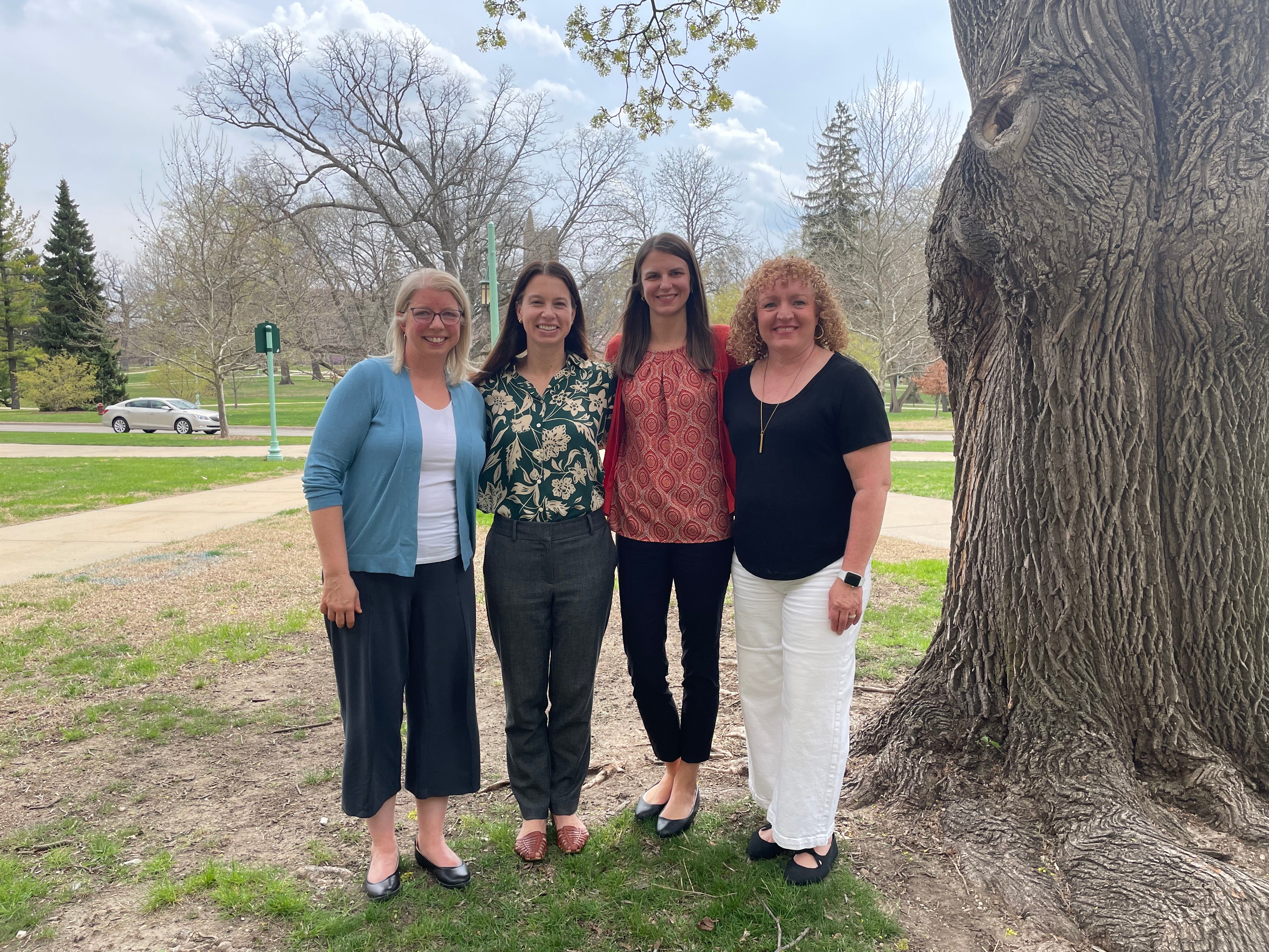 Faculty smile as a group next to a large tree on campus.