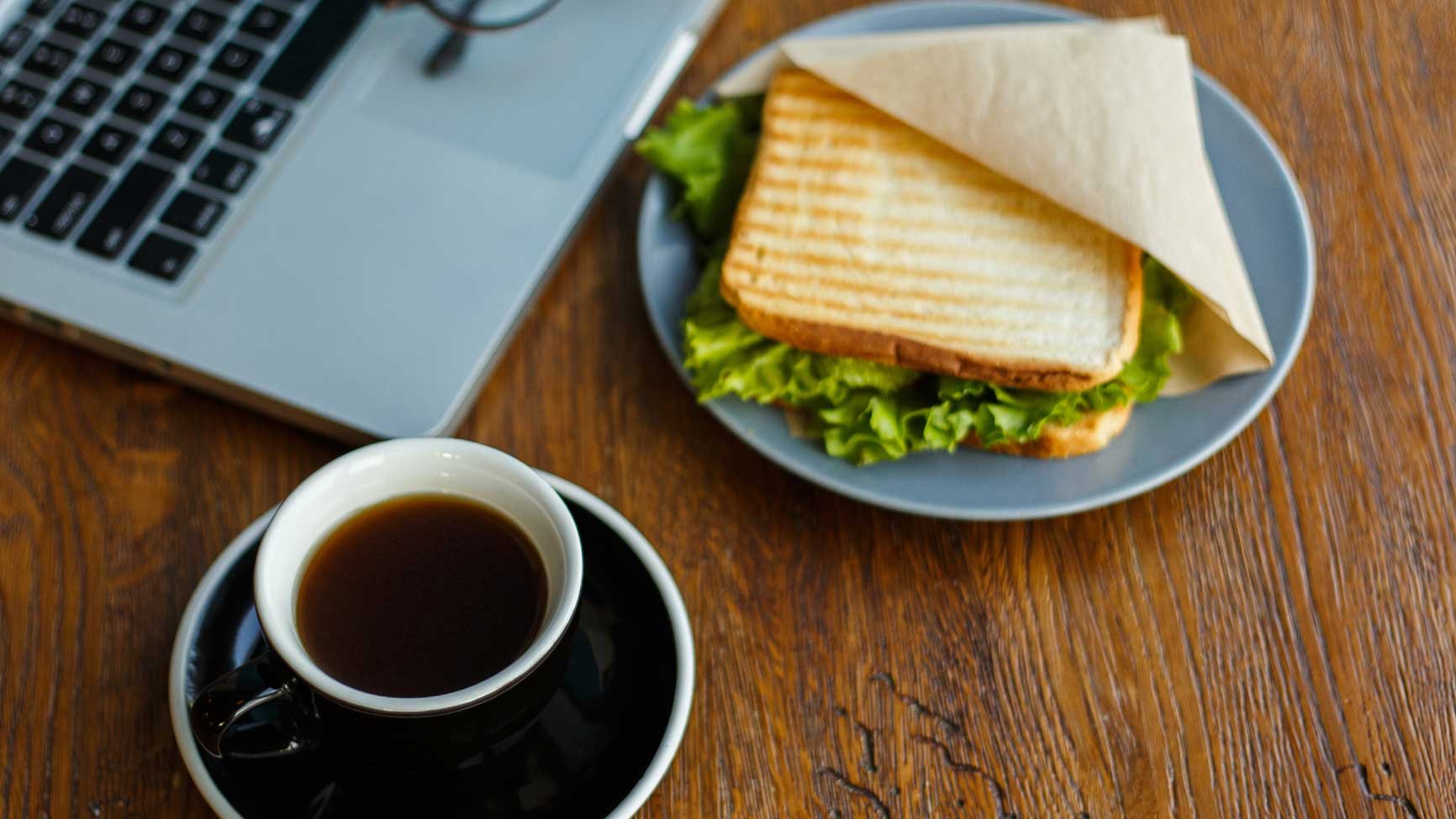 A sandwich and cup of coffee on a desk near a computer.