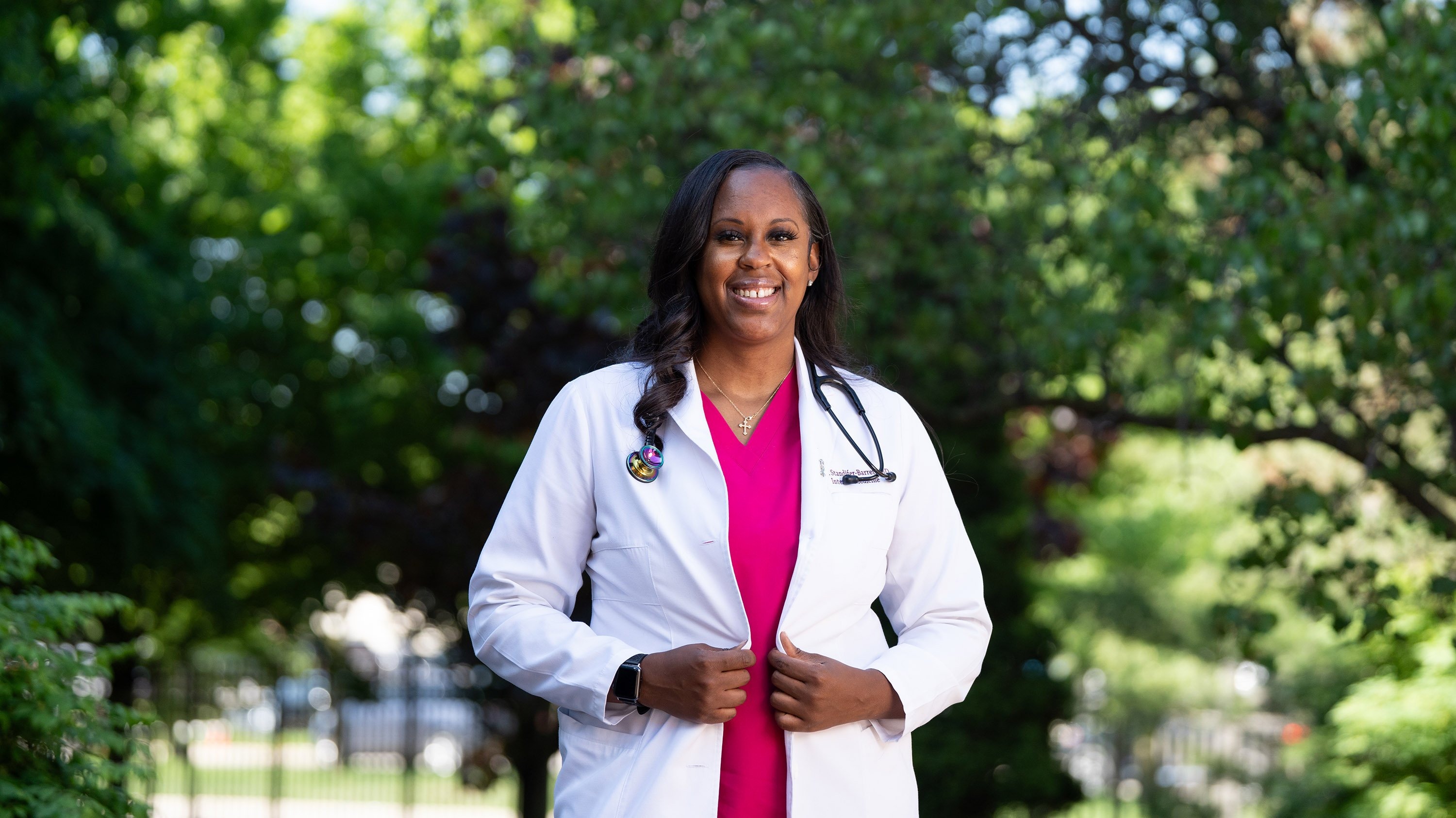 Standifer-Barrett smiling and posing in front of trees wearing her white coat and a pink shirt.