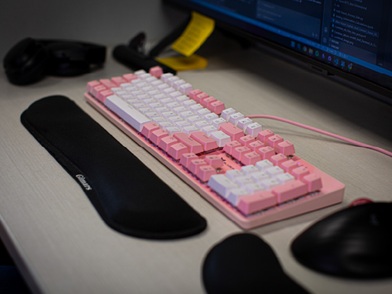 Pink and white keyboard on a desk with a mouse, monitor, and other accessories.