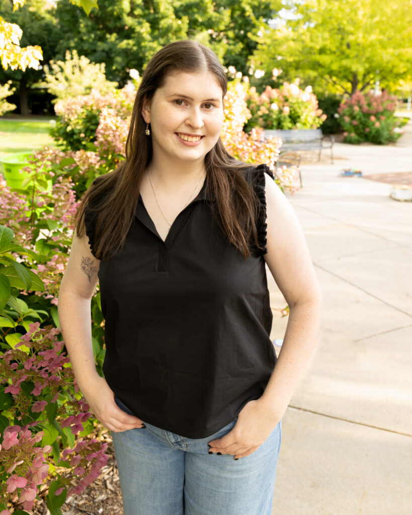 Full-length outdoor portrait of Catelyn Arnold standing on campus walkway next to blooming flowers.