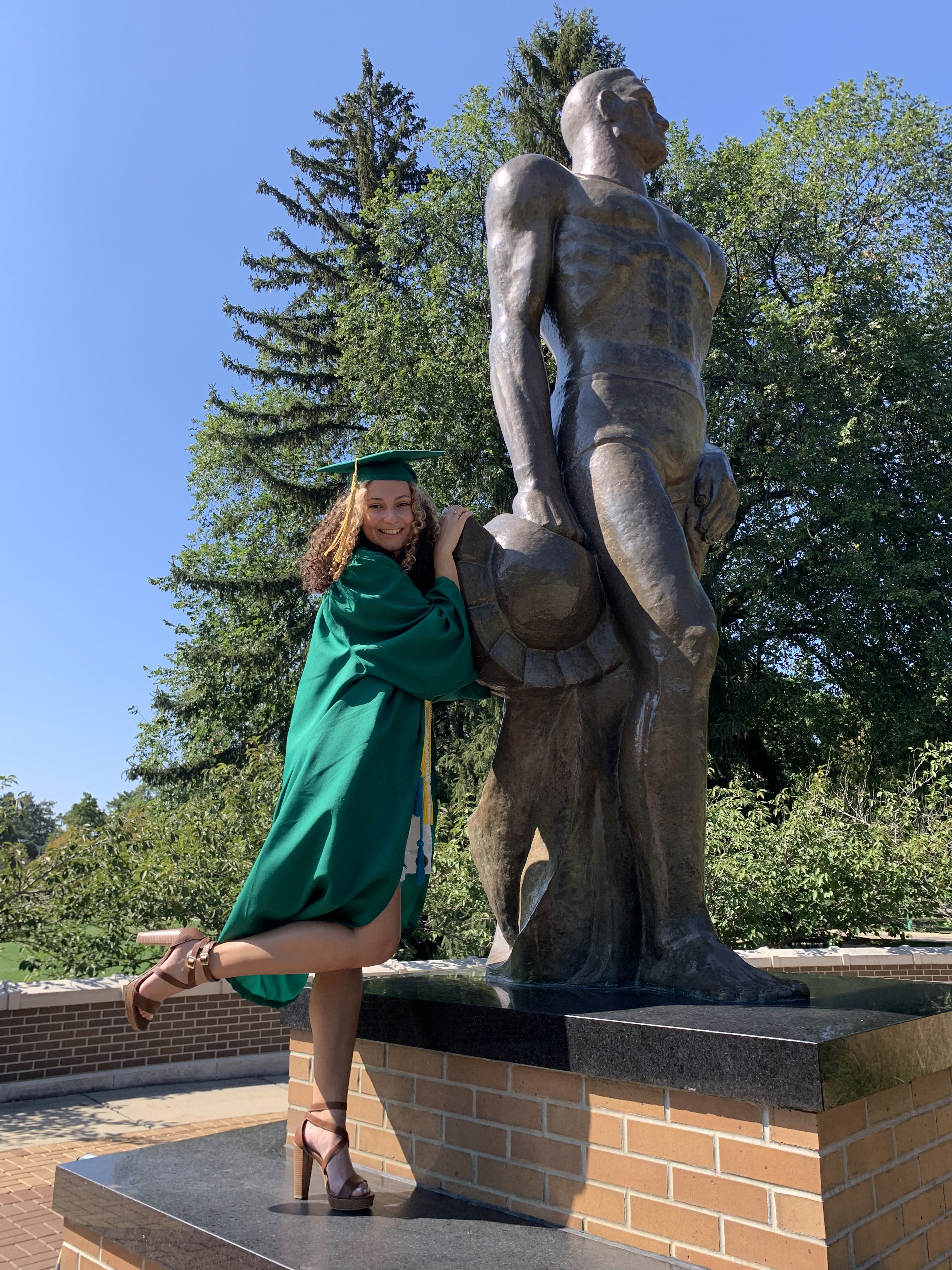 Global and International Studies alumnae Grace Provenzano in her MSU green cap and gown, standing next to the Sparty statue.