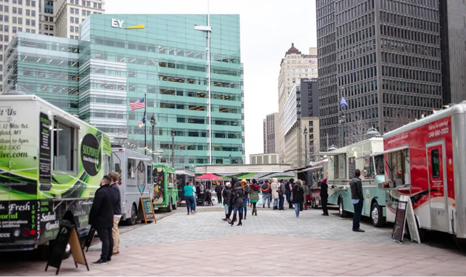 Several food trucks line a city plaza as people gather and walk between them, with high-rise buildings in the background.