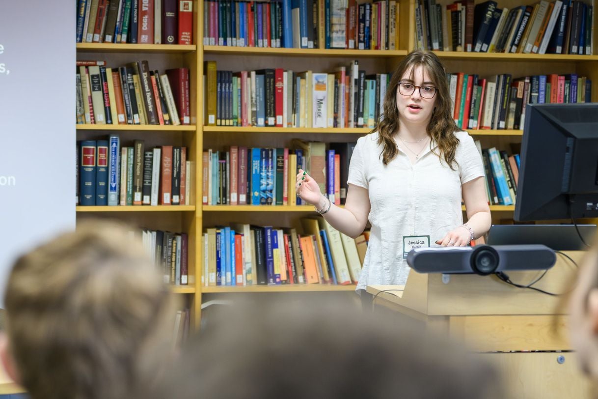 A student in a white blouse presents in front of a wall of books in the JMC Library.
