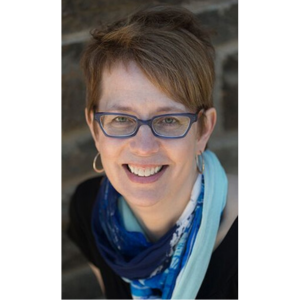 JMC Prof. Allison Berg, photographed outside in front of a brick background. She is wearing blue glasses and a blue patterned scarf.