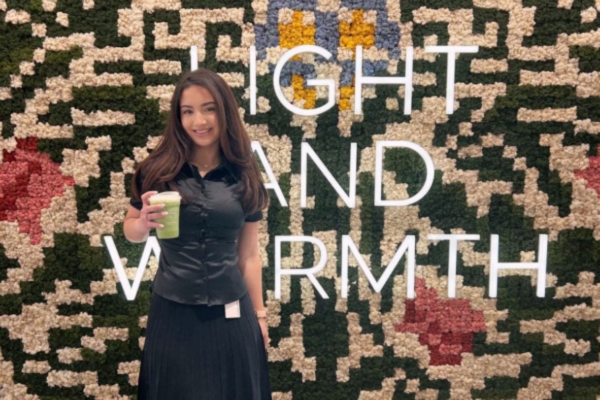 Elena standing in front of tapestry that reads "Light and Warmth" holding a coffee.