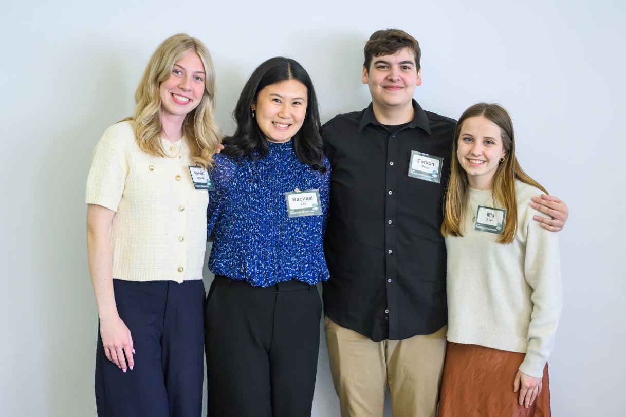 Four JMC students stand together against a white wall, smiling at the camera with arms around each other. All wear Research Showcase nametags.