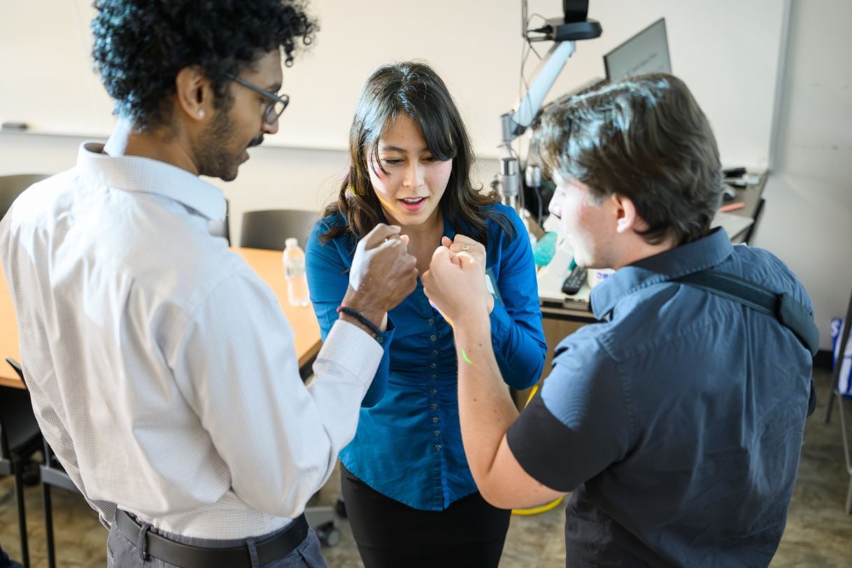 Three students engaged in animated discussion, with the student in the center gesturing emphatically with both hands while two others lean in to listen.