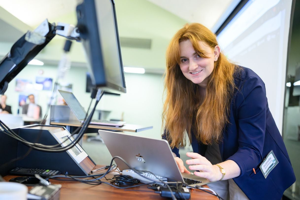 A student in a navy blazer leans over a laptop at the presenter's station, smiling at the camera while preparing her materials.