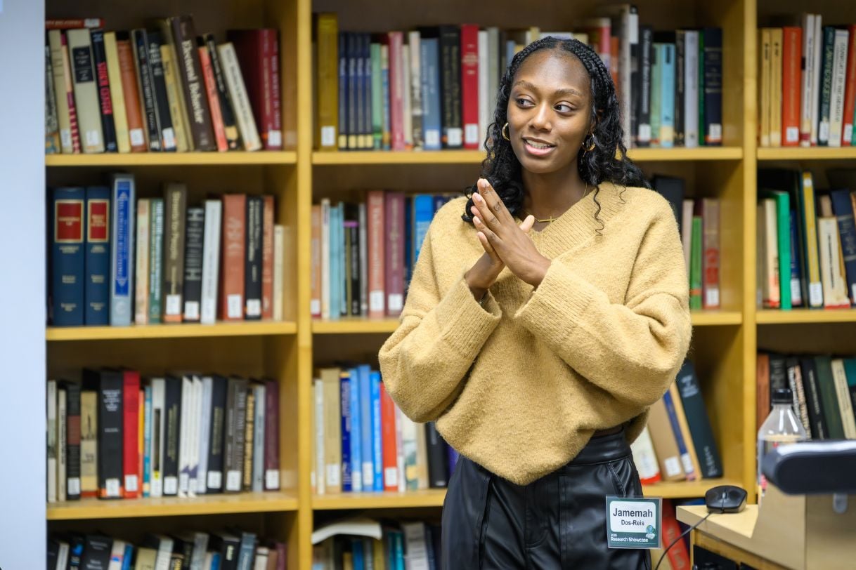 A student in a tan sweater gestures mid-sentence while presenting, framed by floor-to-ceiling shelves of books.