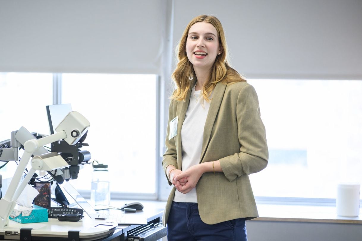 A student in an olive blazer smiles at the camera while standing beside presentation equipment in a research showcase setting.