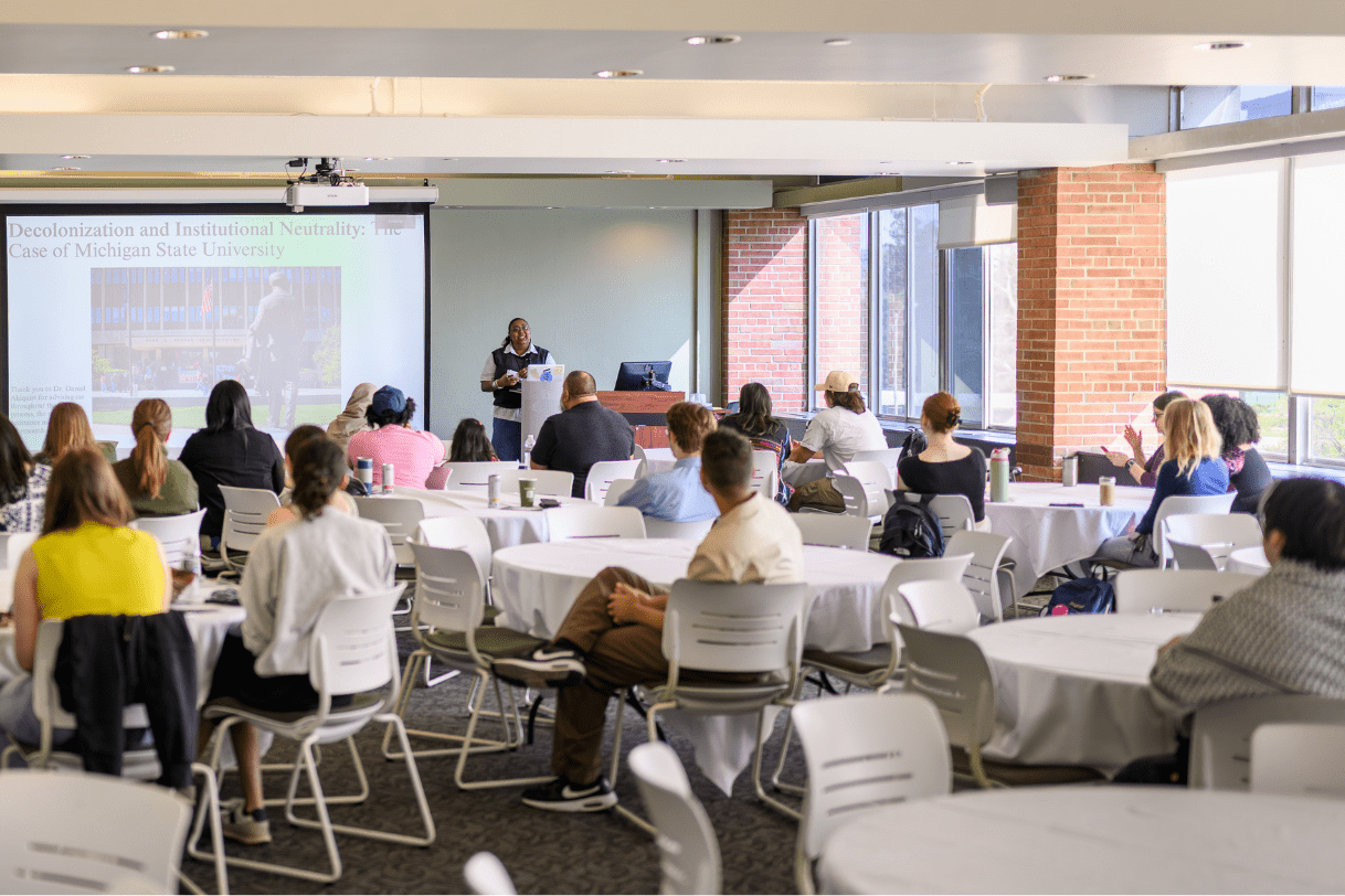 A Madison student presenting during the annual Research Showcase in Club Spartan. The study is shown standing in front of a large screen with an audience of 20-30 people in front.