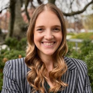 A JMC staff member with long, wavy blonde hair smiles warmly at the camera. She is wearing a dark, vertically striped blazer and stands outdoors against a backdrop of trees and greenery.