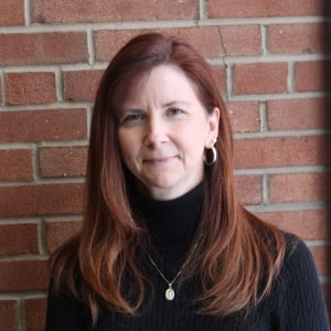 A JMC staff member with long red hair stands against a brick wall, wearing a black turtleneck and small hoop earrings. She looks directly at the camera with a neutral expression.