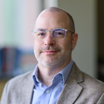 Russell Lucas wearing a tan blazer, light blue collared shirt and glasses.