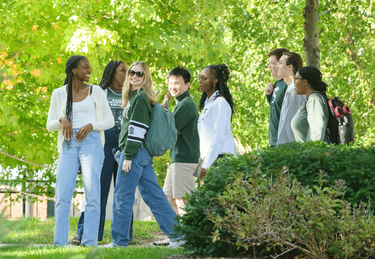 A group of James Madison College students walk together along a grassy path surrounded by vibrant green trees and bushes. They are casually dressed, some carrying backpacks, with expressions suggesting camaraderie as they move through the sunny outdoor setting.