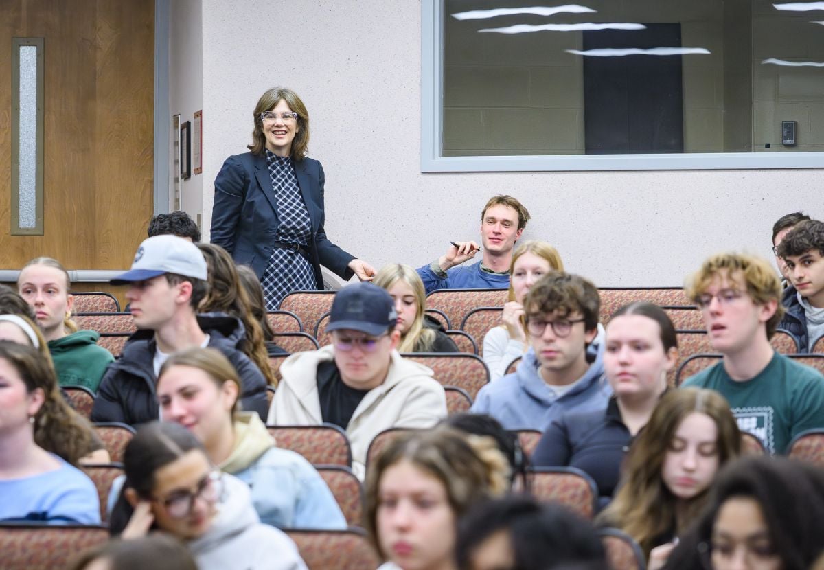 A JMC professor in a dark blazer and patterned blouse stands among rows of seated students, smiling as she appears to address the group. The setting is an indoor lecture hall with red upholstered seats, wooden doors, and large windows.