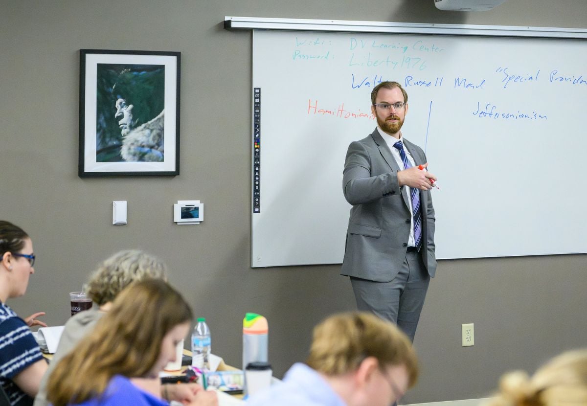 A JMC professor with glasses and a beard, dressed in a gray suit, stands at the front of a classroom holding a red marker while writing on a whiteboard. Several students are seated at desks, focused on their work or listening to his lecture.