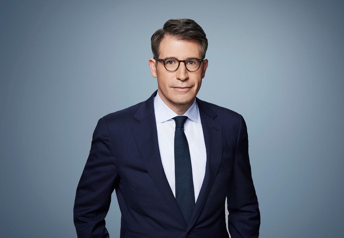 A JMC alum wearing a dark navy suit, white shirt and tie. He is wearing glasses and smiling at the camera, against a blue backdrop.