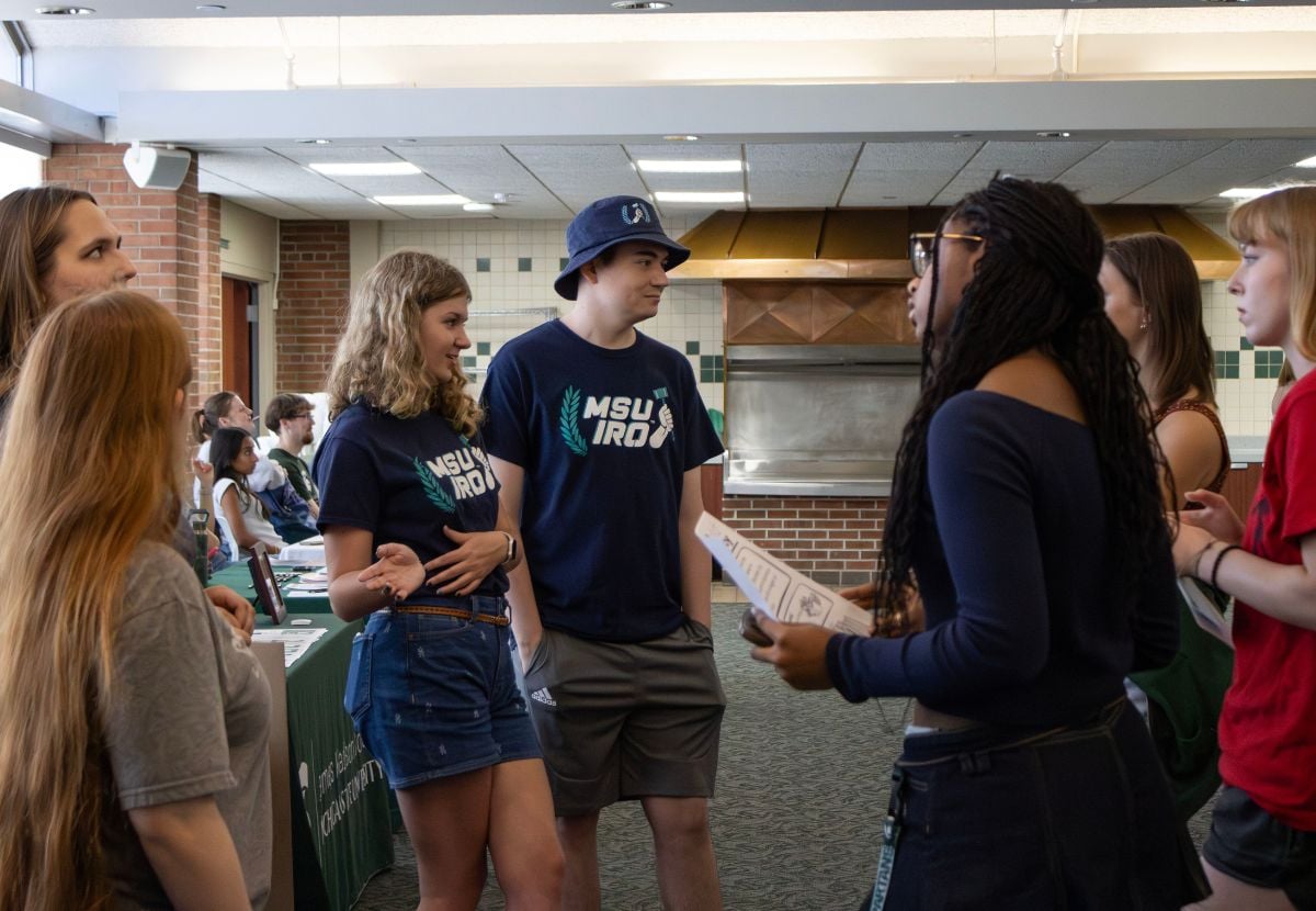 A group of JMC students, including two wearing MSU IRO t-shirts, are interacting and conversing in what appears to be a campus event or information session. The setting includes tables with materials, other attendees, and indoor architectural features like brick walls and ceiling lights