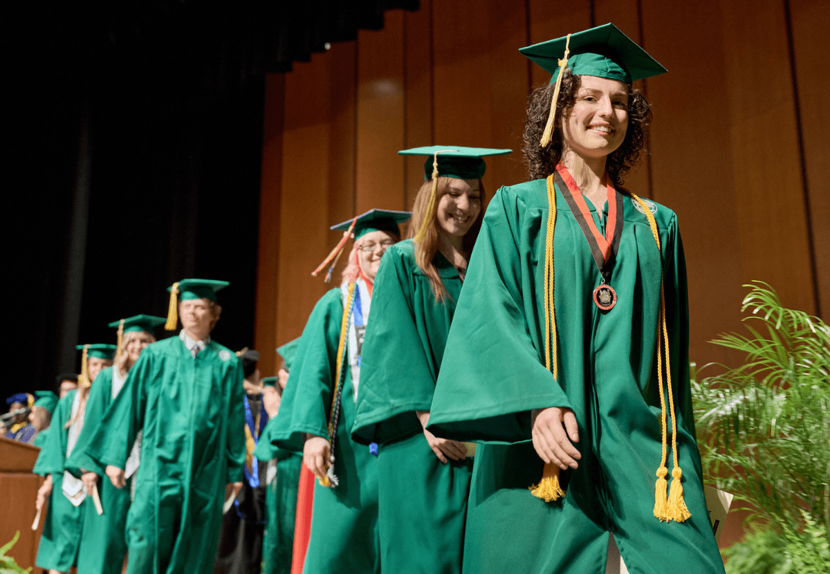 Image shows JMC students wearing green graduation gowns and caps walking across a stage. 