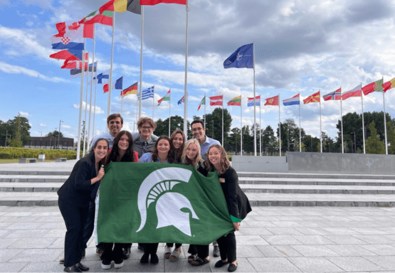 Photo shows a group of Madison students posing with a green MSU flag with the Spartan helmet on it.