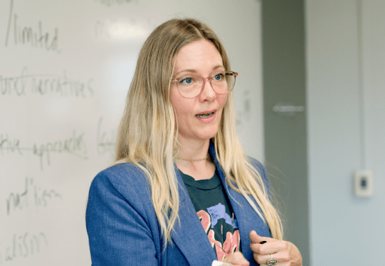 Assoc. Prof. Amanda Flaim wearing a blue blazer and glasses while teaching in front of a white board.