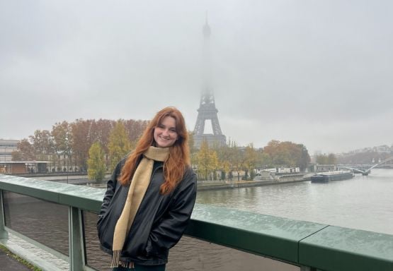 Photo shows JMC student Isabelle Radkovich posing in front of the Eiffel Tower (from a distance) on a cloudy day. She is wearing a black jacket and gold scarf. 