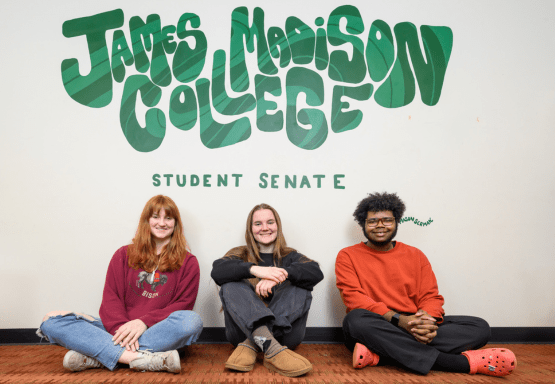 Three Madison students sitting cross-legged on the floor in front of a mural that reads James Madison College Student Senate.