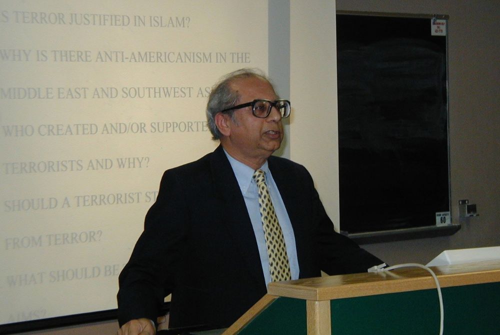 A man wearing glasses and a suit stands behind a podium, speaking to an audience. Behind him, a projection screen displays questions.