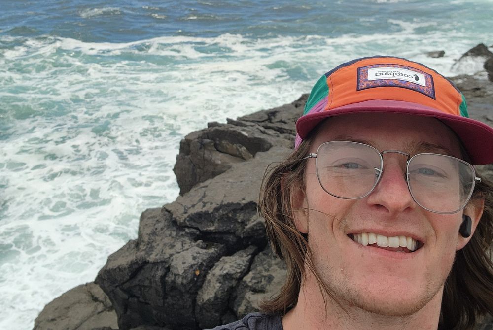 JMC student Colin Reese photographed outside on the coast of Ireland. He is wearing glasses and an orange cap.