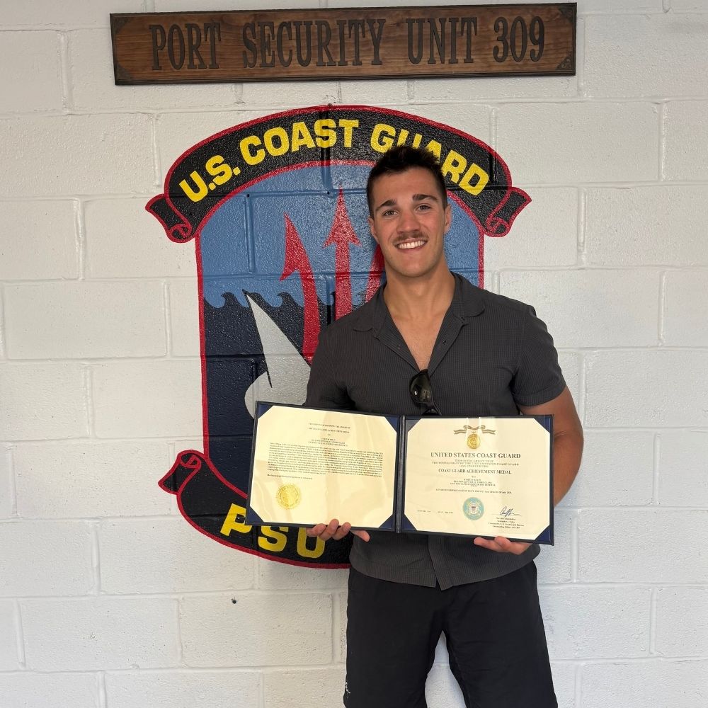 A JMC student stands in front of a U.S. Coast Guard emblem, holding two framed certificates. He is wearing casual attire and appears to be celebrating his achievement at Port Security Unit 309.