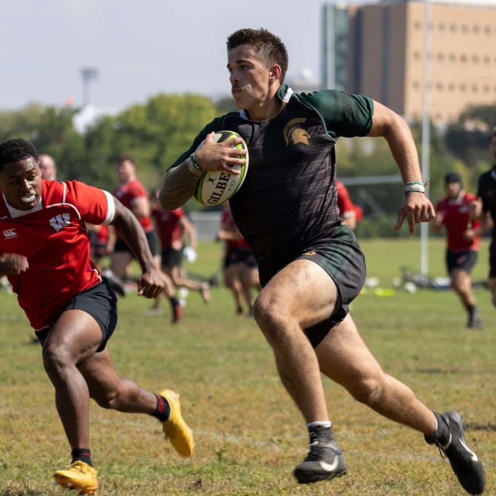 A JMC student who is a rugby player in a green and black uniform sprints across the field while holding a Gilbert rugby ball. Another player in red runs alongside him, attempting to tackle or intercept.