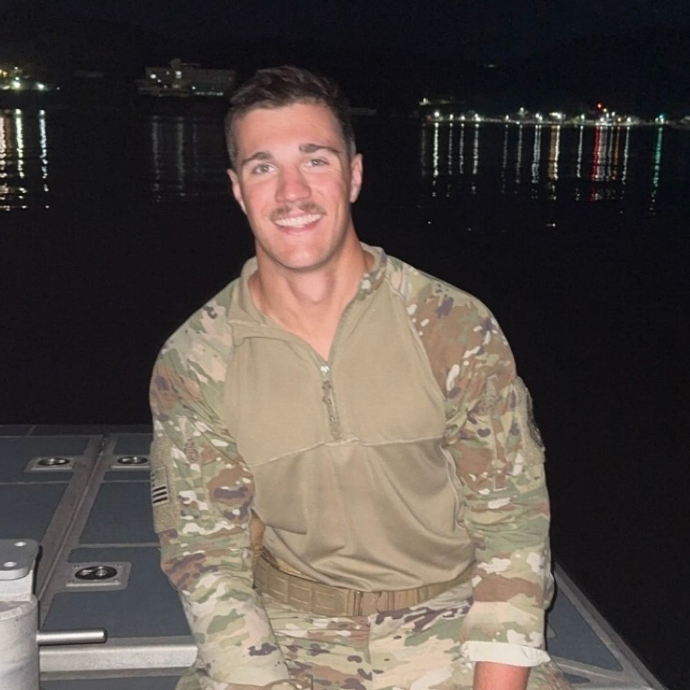 A JMC student who is a part of the Coast Guard in camouflage uniform smiles at the camera while seated on a boat. The background shows dark water with distant city lights reflecting on its surface.