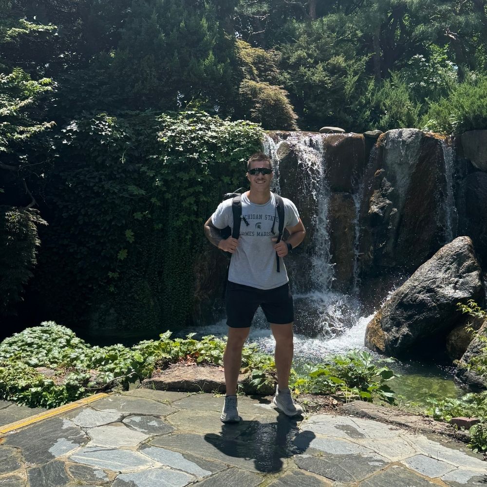A JMC student wearing sunglasses and a backpack stands on a stone path in front of a small waterfall surrounded by lush greenery. He is smiling at the camera while holding his backpack straps.