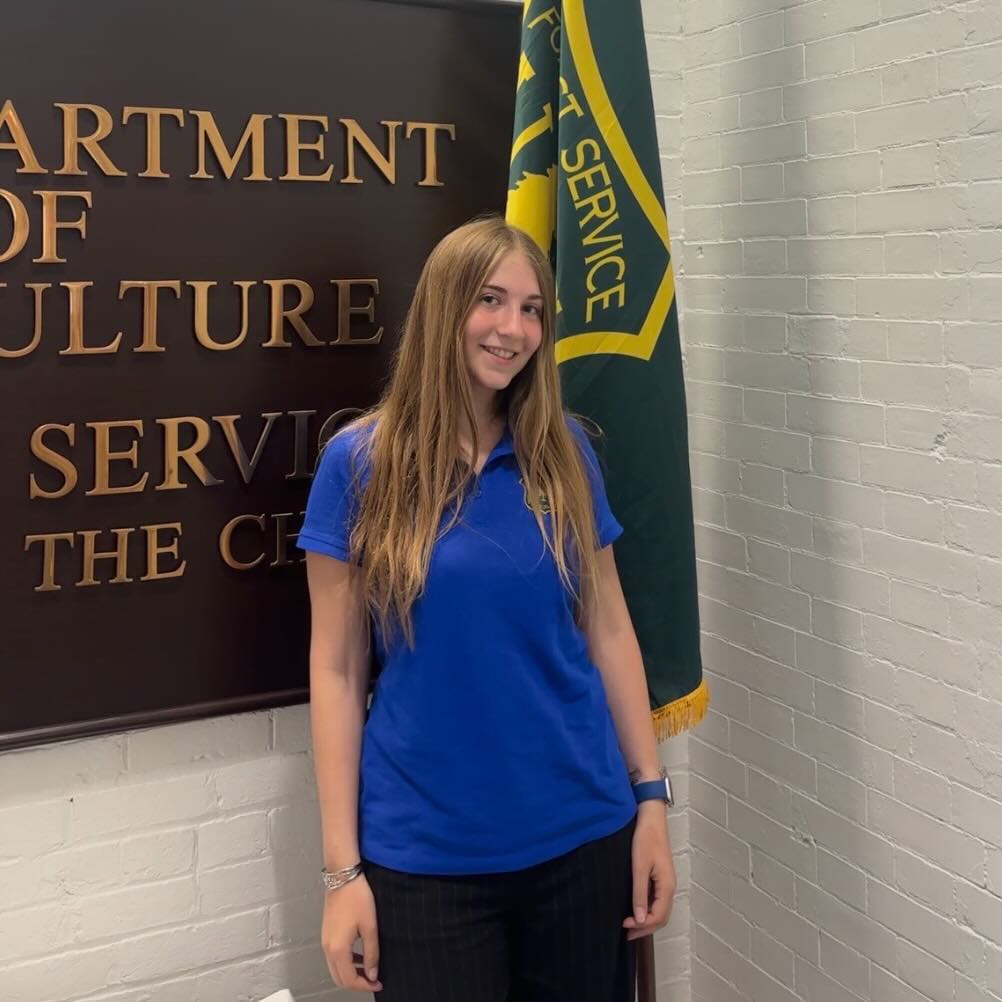 A JMC student photographed in front of a U.S. Forest Department plaque and flag. The student is wearing a blue polo shirt.