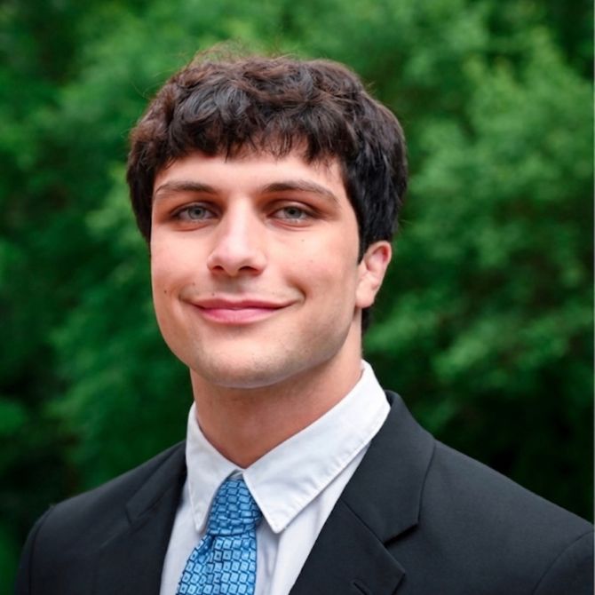 A JMC student with dark hair and blue eyes is smiling while wearing a black suit, white shirt, and patterned tie. He stands outdoors against a blurred green background of trees.