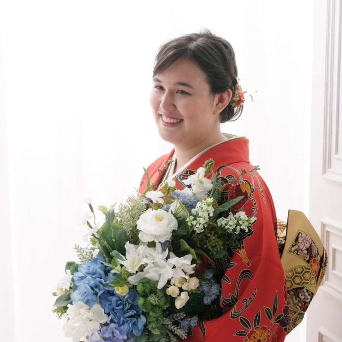 A JMC student wearing a vibrant red kimono with intricate floral patterns smiles while holding a large bouquet of white and blue flowers. She stands indoors near a bright window, her dark hair styled up with decorative accents.