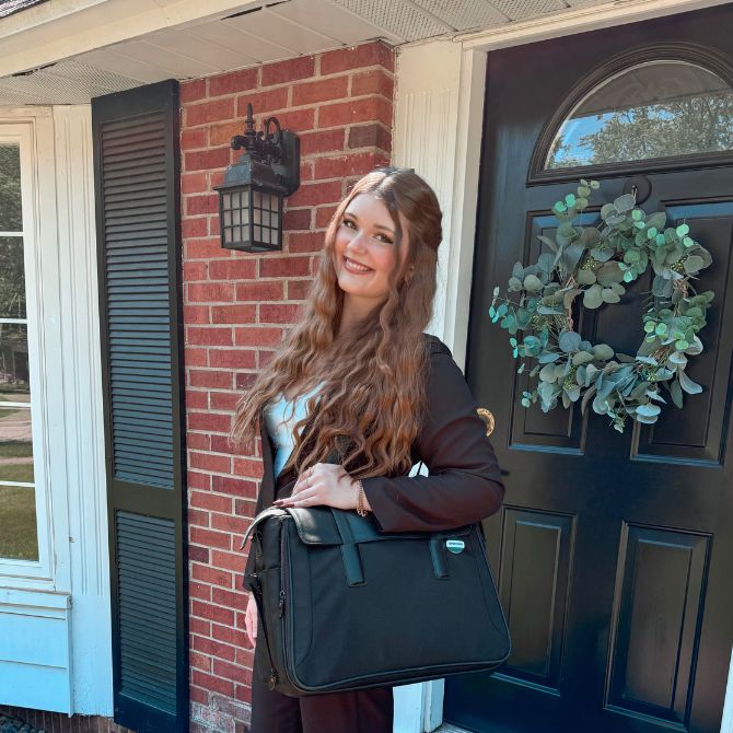 A JMC student with long, wavy hair stands smiling in front of a brick house, holding a black briefcase. She is positioned beside a dark wooden door adorned with an eucalyptus wreath, under the shade of a porch overhang.