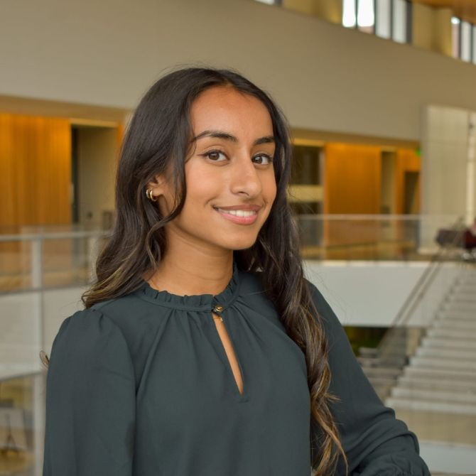 A JMC student with long dark hair smiles warmly at the camera while standing indoors. She is wearing a dark green blouse and small hoop earrings, positioned near glass railings within a spacious, modern architectural space.