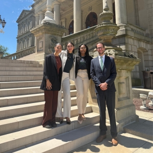 Four people standing together in professional dress, outdoors on steps in front of the Michigan Capitol building.
