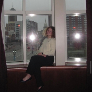 A JMC student photographed sitting on an indoor window ledge, with the capitol building visible in the view behind.