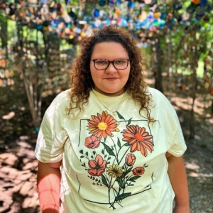 A JMC student photographed outdoors, wearing a bright yellow shirt with flowers on the front. The student is wearing glasses and smiling at the camera on a sunny day.