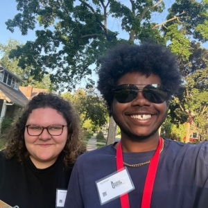 Two JMC students outdoors taking a selfie. The student on the left has curly hair and glasses. The student on the right is wearing sunglasses and a red lanyard.