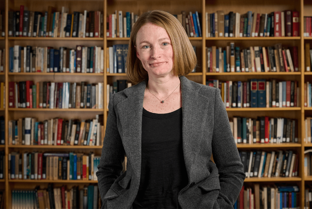 JMC Prof. Kirstin J.H. Brathwaite in the college's library. She is wearing a grey blazer and posing front of a wall of bookshelves.