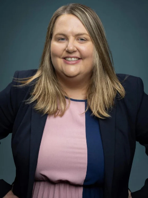 A JMC alumna smiling at the camera in a professional portrait. She is wearing a blue blazer and a pink shirt. 