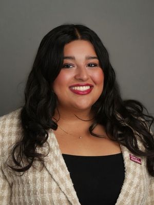 A JMC student with long dark hair smiles warmly at the camera. She wears a beige blazer over a black top, standing against a plain gray background.