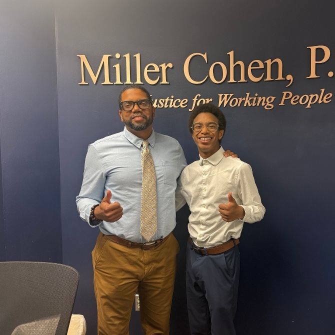 Two men stand smiling and giving thumbs up in front of a blue wall with the sign 'Miller Cohen, Justice for Working People'. The man on the left wears glasses, a light blue shirt, patterned tie, and khaki pants, while the younger man on the right wears glasses, a white button-up, and dark pants.