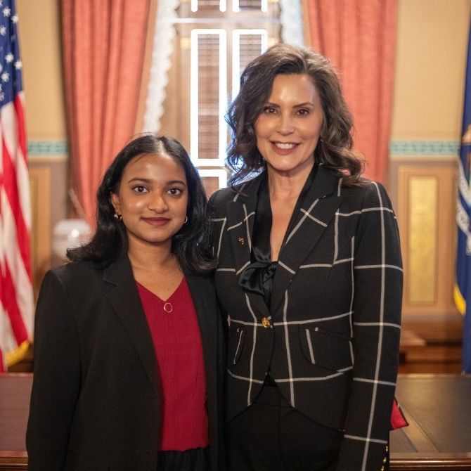 JMC student standing next to Michigan Governor Gretchen Whitmer. Student is wearing a red shirt and black blazer. The State of Michigan and American flags are visible in the background.