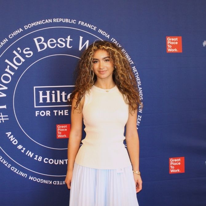 A JMC student with curly hair stands confidently in front of a blue backdrop featuring the "World's Best Workplace" logo and "Hilton" branding. She is wearing an elegant white sleeveless top and light-colored skirt, posing for the camera.